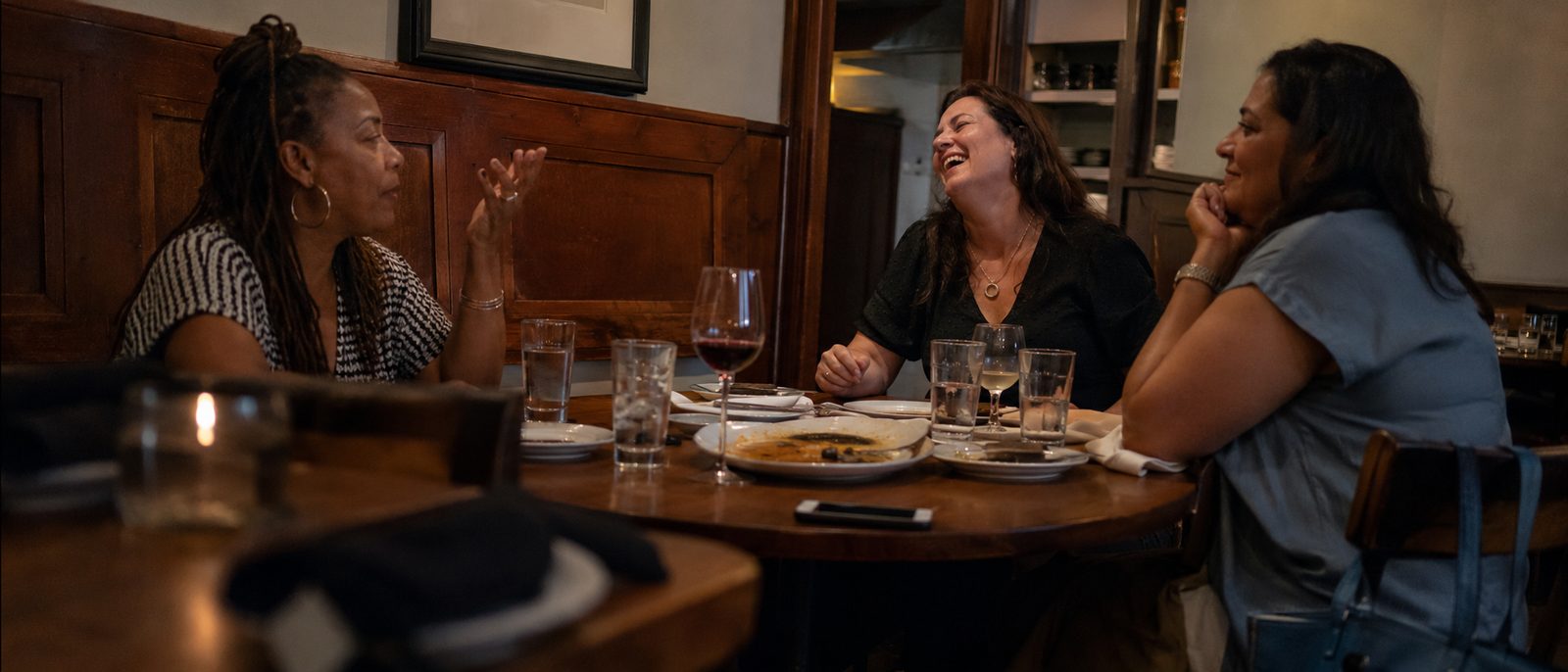 Three women between forty and fifty-five at a restaurant table, one mid-sentence, one laughing.