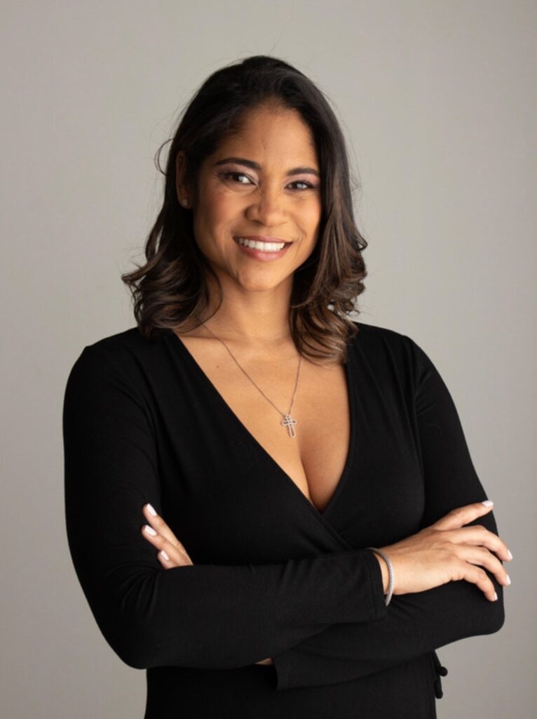 Cathy Goodall-Jackson, arms folded, smiling at the camera against a plain warm grey background.
