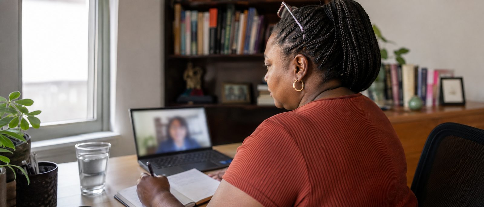 A woman at a laptop in her home office during a video consultation, writing questions in a notebook.